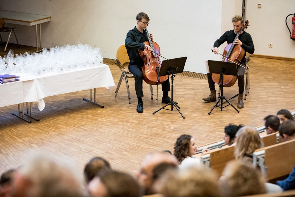 Blick aus dem Publikum, vorn im Bild unscharf Hinterköpfe, dann zwei Cellisten auf der Bühne. Rechts neben ihnen der Tisch mit den Urkunden und Geschenken.