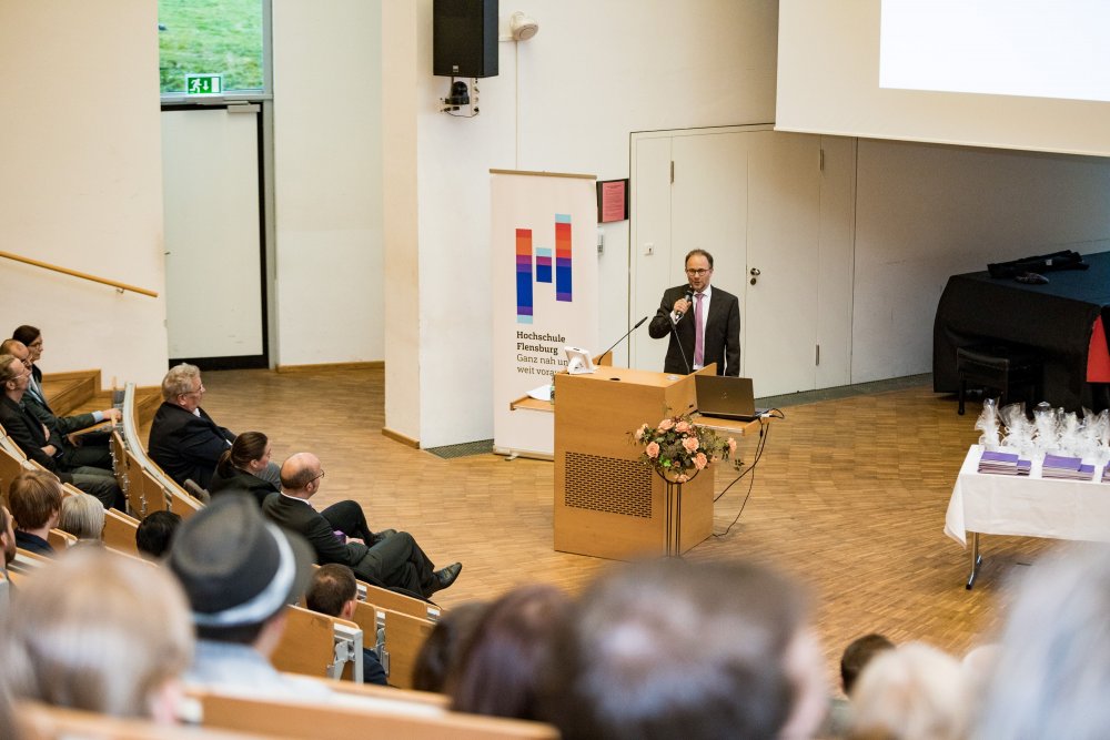 Sehr klein im Bild ist der Gatredner am Rednerpult, im linken Bildrand Hinterköpfe des Publikums.