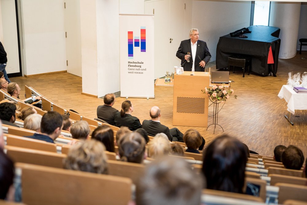 Der Gastredner gut zu erkennen im Fokus, er hält ein Bierglas in der Hand, außerdem Hinterköpfe des Publikums.