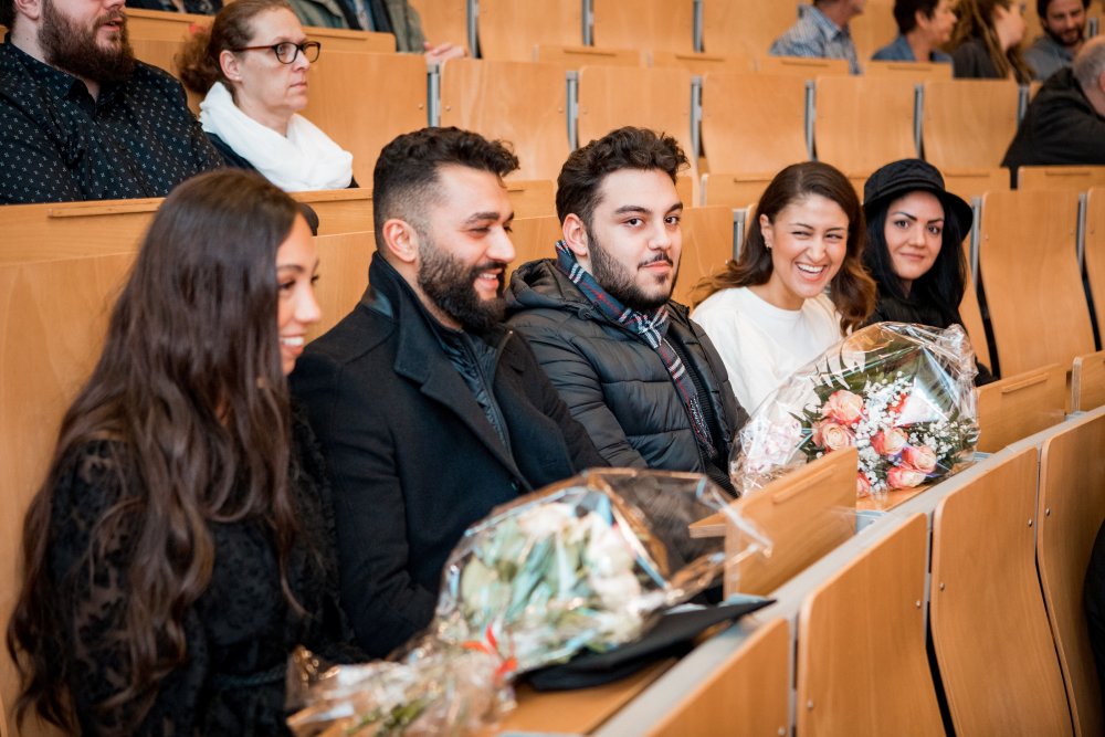 Fünf junge Menschen sitzen in einer Reihe in einem hörsaalartigen Raum. Sie sind elegant gekleidet und lachen.