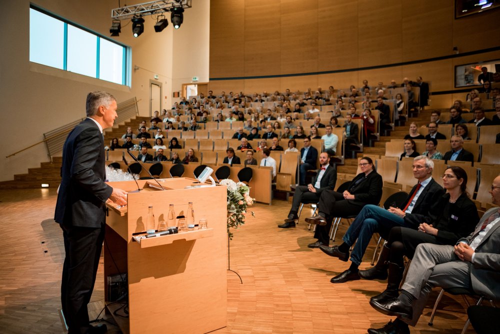 Links im Bild schräg von hinten zu sehen: Ein Redner im Anzug an einem Pult. Rechts, also vor ihm, das Publikum.