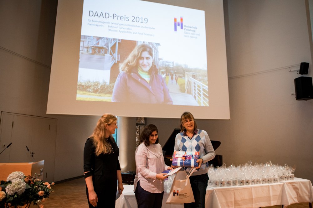 Drei Frauen stehen auf der Bühne, Die Frau in der Mitte bekommt ekine Urkunde überreicht. Sie ist auch auf einem Bild, das an die Leinwnad hinter ihr projeziert wird, zu erkennen. Darüber steht "DAAD Preis".
