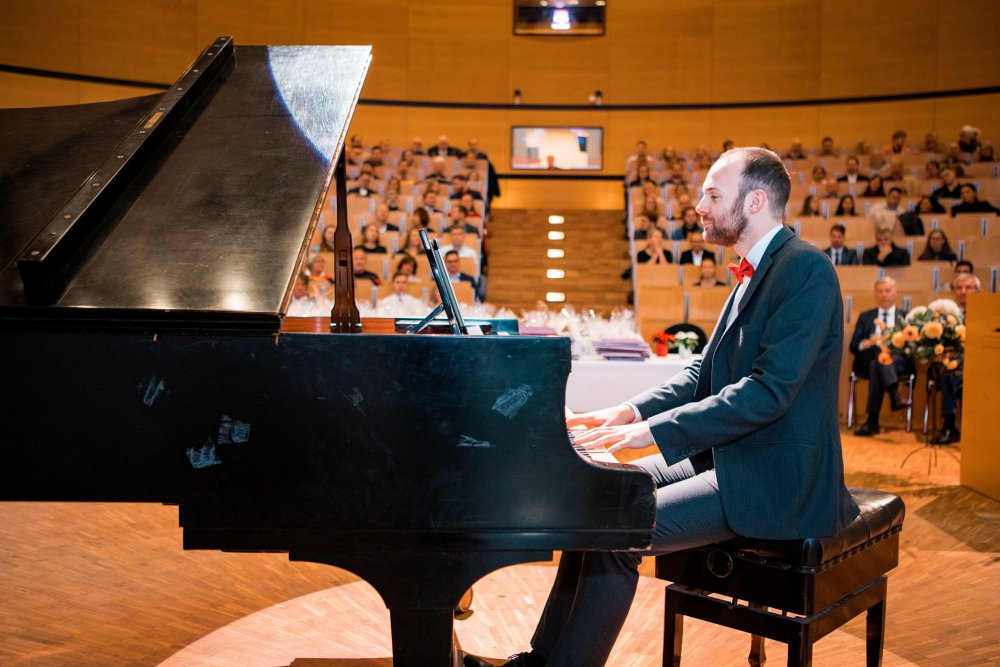 Ein Klavierspieler in Anzug. Dahinter ist das Publikum unscharf zu erkennen.