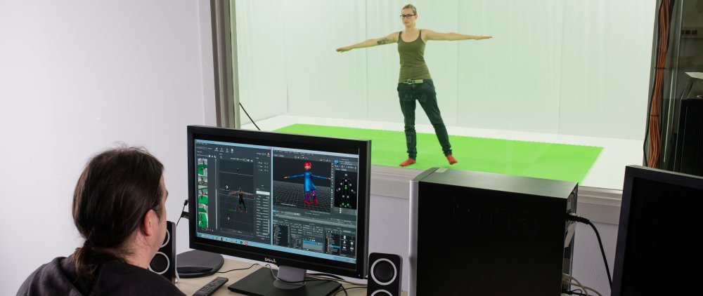 Studentin und Dozent im Motion Capture Studio. Der Dozent sitzt vor der Glaswand an einem Computer, die Studentin steht im ausgebreitetem Armen hinter der Glaswand im Studio.