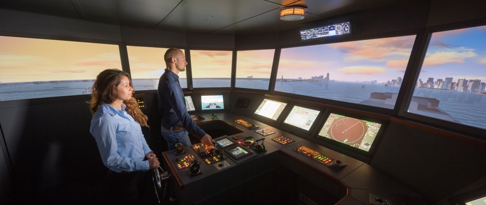 Studierende im Schiffsführungssimulator. Vor ihnen beleuchetete Bedienungspanel, durch die Fenster der Schiffbrücke ist eine Szene auf See zu erkennen.