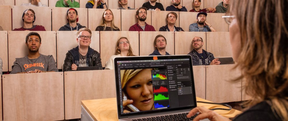 Studierende im Hörsaal. Vorn, eher rechts im Bild und angeschnitten der Hinterkopf einer Dozentin, die einen Laptop bedient.
