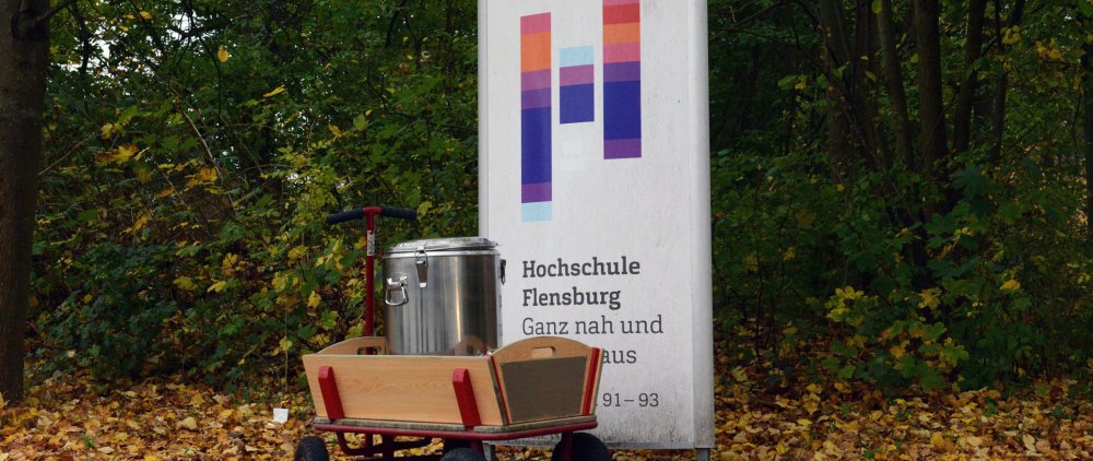 Vor einem Schild mit dem Hochschullogo auf weißem Grund steht ein Bollerwagen mit einem silbernen Thermosfass darauf. Auf dem Boden liegt gelbes und braunes Laub, im Hintergrund sind außerdem Bäume zu erkennen.