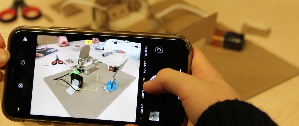 Eine Hand hält ein Handy, auf dem eine 3D Simulation zu sehen ist. Im Hintergrund ein Tisch mit Bastelmaterialien.