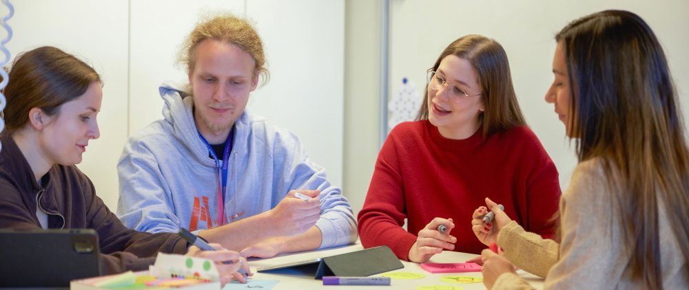 Vier Studierende sitzen um einen Tisch, darauf Arbeitsmaterialien wie bunte Zettel und Stifte. Sie besprechen sich.