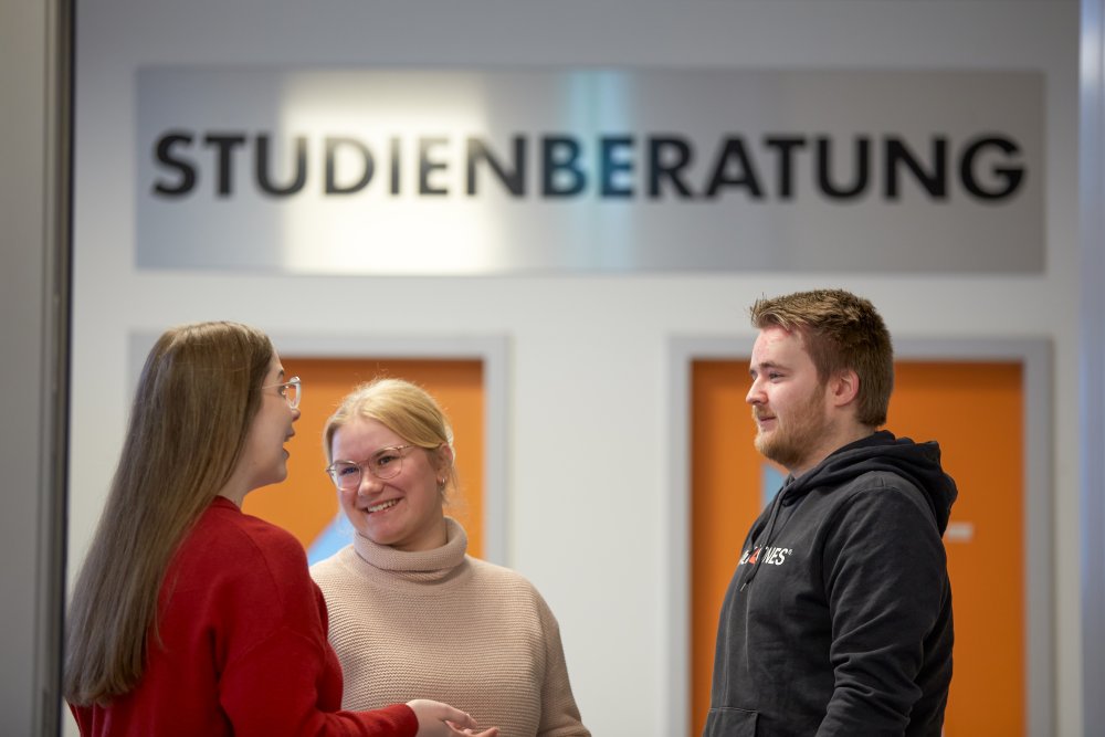 Drei Studierende unterhalten sich lachend. Im Hintergrund sieht man zwei orangene Türen, über denen in metallischen Großbuchstaben das Wort "Studienberatung" steht.