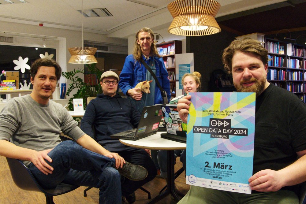 Vier Männer unterschiedlichen Alters und eine junge Frau sitzen rund um einen kleinen runden Tisch, auf dem zwei aufgeklapte laptops stehen. Der Mann im Vordergrund hält ein buntes Plakat mit der Aufschrift "Open Data Day 2024 - Flensburg, 2. März" in die Kamera.