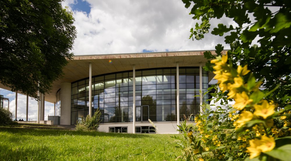 Das Audimax der Hochschule Flensburg: Ein modernes Gebäude mit Glasfassade und dreieckigem Flachdach, im Vordergrund eine grüne Wiese, Büsche und leuchtend gelbe Blüten.
