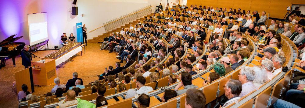 In einem großen Hörsaal mit voll besetzten Rängen steht ein Mann vorne am Pult und spricht zum Publikum, neben ihm ein Tisch mit dekorativ aufgebauten Präsenten.
