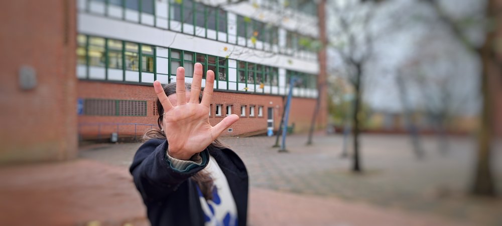 Eine Frau steht auf einem Hof zwischen zwei Backsteingebäuden. Sie hat die Hand abwehrend vor sich ausgestreckt, sodass man ihr Gesicht nicht erkennt.