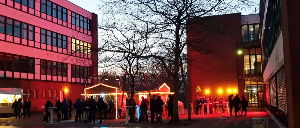 Auf einem Platz zwischen zwei großen Backsteingebäuden stehen weihnachtlich beleuchtete Buden, an Stehtischen stehen zahlreiche Menschen in Grüppchen und unterhalten sich, es ist Abend.