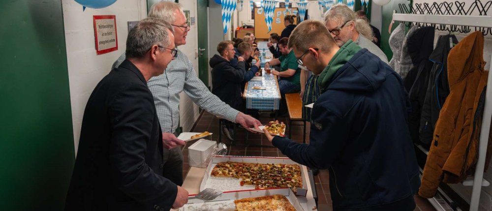 An einem Biertisch stehen zwei Männer und teilen Pizza aus, im Hintergrund stehen weitere Biertisch-Garnituren, an denen zahlreiche junge Leute sitzen und sich unterhalten.