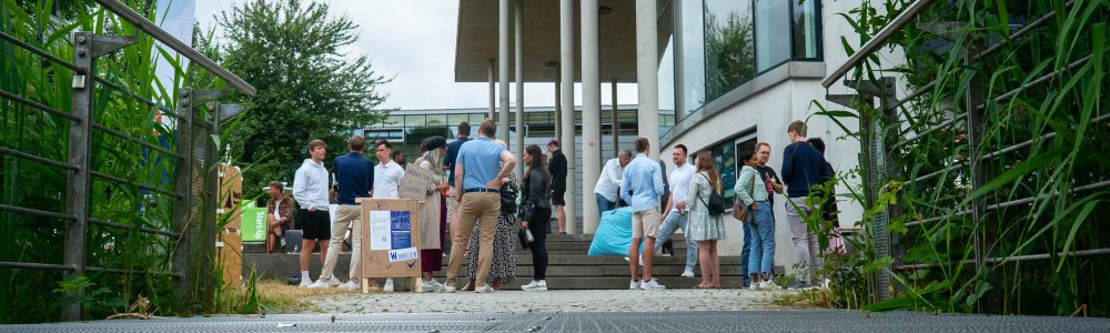 Mehrere Grüppchen von Menschen verschiedenen Alters stehen neben einem modernen Gebäude mit Säulen auf und an einer Treppe und unterhalten sich angeregt.