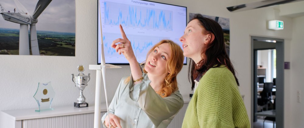 Two women pointing and looking upwards at the tip of a wind turbine model.