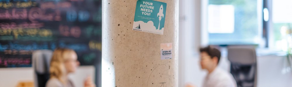 Auf einer weißen Säule klebt ein Sticker mit der Aufschrift "Your future needs you!", im Hintergrund sieht man verschwommen zwei Personen an Schreibtischen und eine bunt beschriebene Tafel.