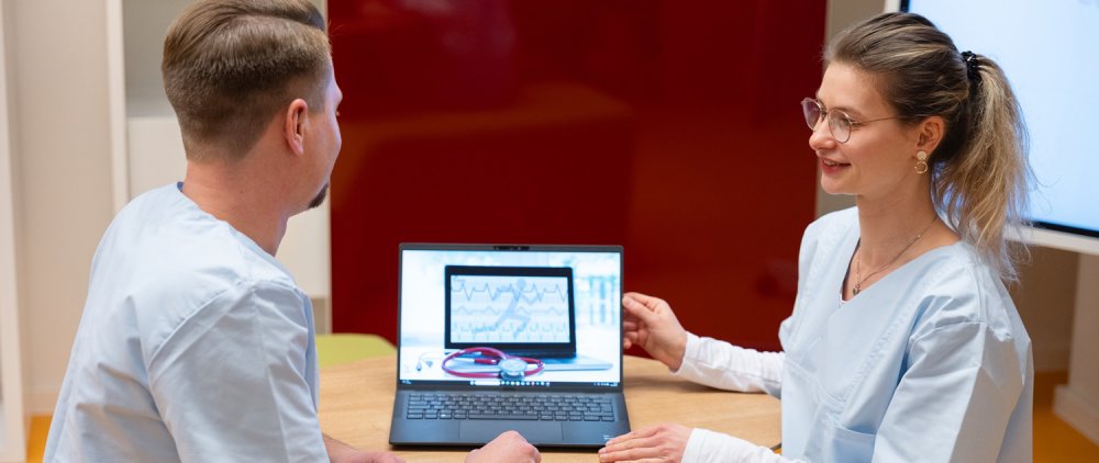 Eine junge Frau und ein junger Mann in hellblauen Kitteln blicken gemeinsam auf einen Laptop.
