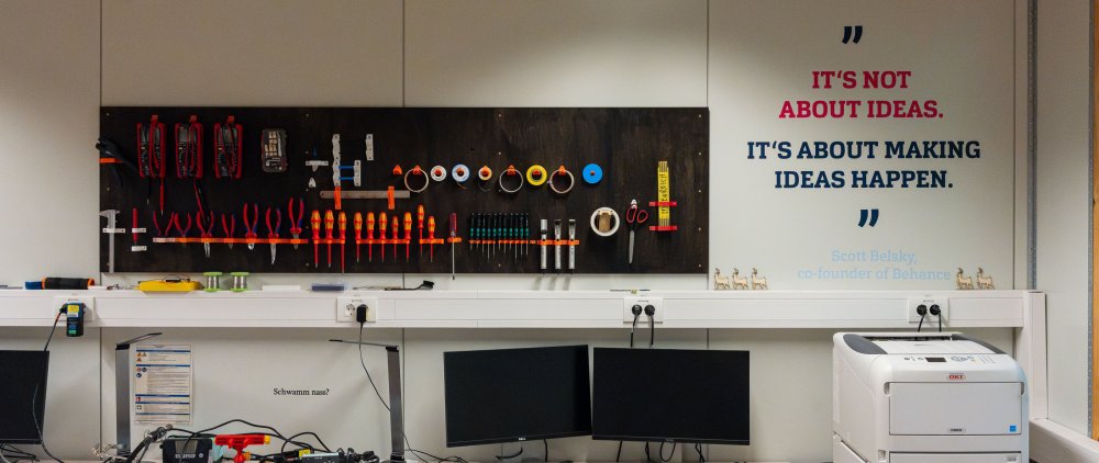 Wand, an der Werkzeug hängt. davor Bildschirme und ein Drucker. An der Wand steht in englischer Sprache: ItÄs not about ideas. It's about making ideas happen.