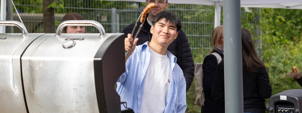 Ein junger Mann steht hinter einem Grill und hält mit einer Grillzange lachend eine Wurst hoch.