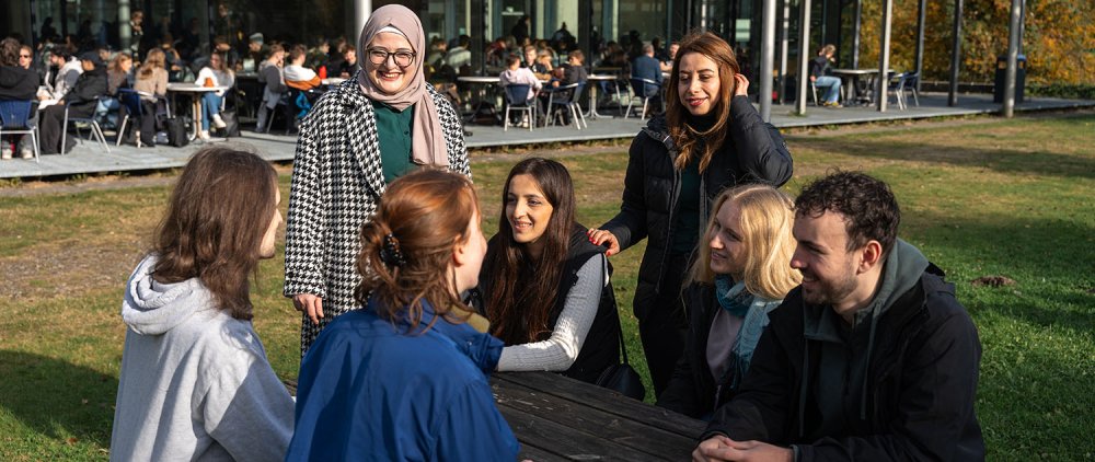 Eine Gruppe von Studierenden sitzt an einem Picknicktisch hinter der Mensa Flensburg und unterhält sich angeregt.
