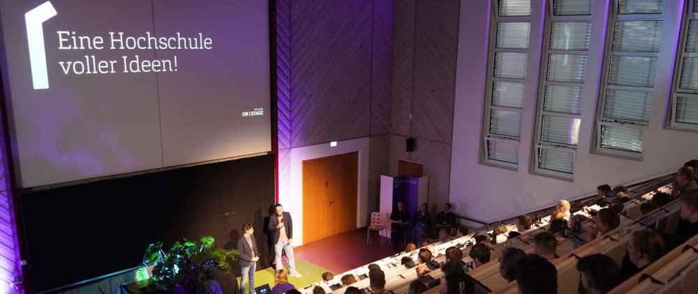 In einem gut gefüllten Hörsaal stehen zwei Männer vorne im Scheinwerferlicht und sprechen zum Publikum, über ihnen eine Projektion mit Text: Eine Hochschule voller Ideen!