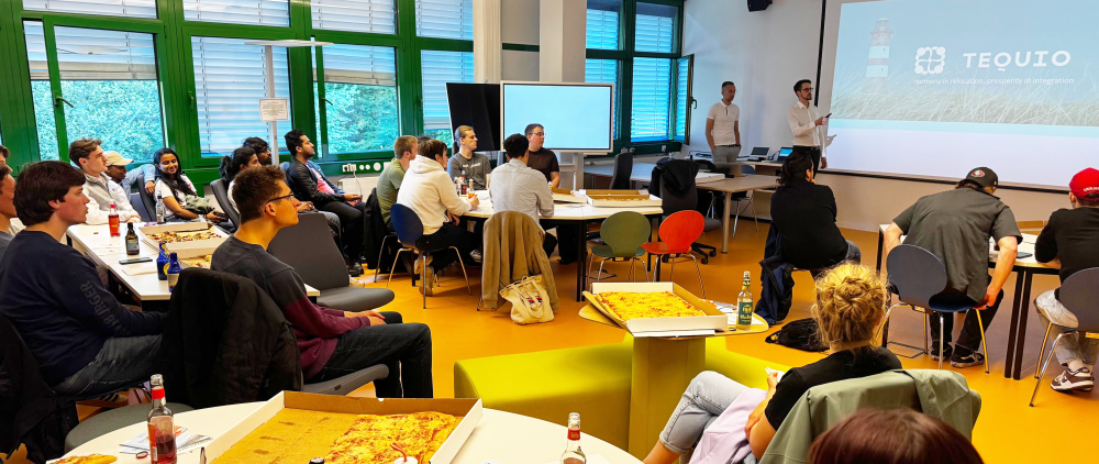 Ca. 30 Studierende sitzen in einem Raum an mehreren runden Tischen, vorne halten zwei eine Präsentation, auf den Tischen stehen Getränke und Partypizzen.