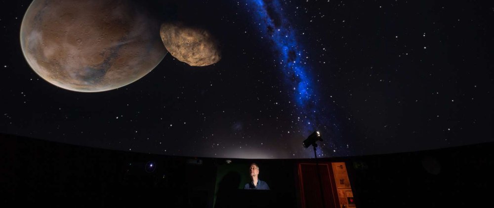Bild einer Planetariumskuppel mit Projektion der Milchstraße und zweier Planeten, darunter steht ein Mann an einem Rednerpult.