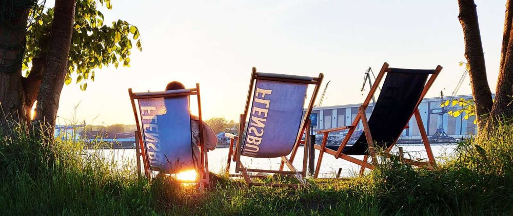 Drei Liegestühle der Marke Flensburger stehen mit Blick auf Sonnenuntergang am Wasser, in einem sitzt eine junge Frau.