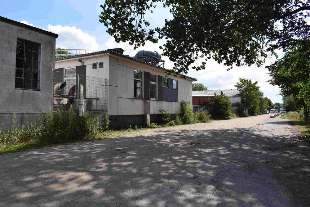 Schmucklose eingeschossige Zweckbauten, davor ein Weg. Der Gimmel ist blau und sonnig. Bäume und Gebüsch am Bildrand und zwischen den Gebäuden sind grün. Zwischen den Gebäuden sind außerdem große Abzugrohre zu erkennen.