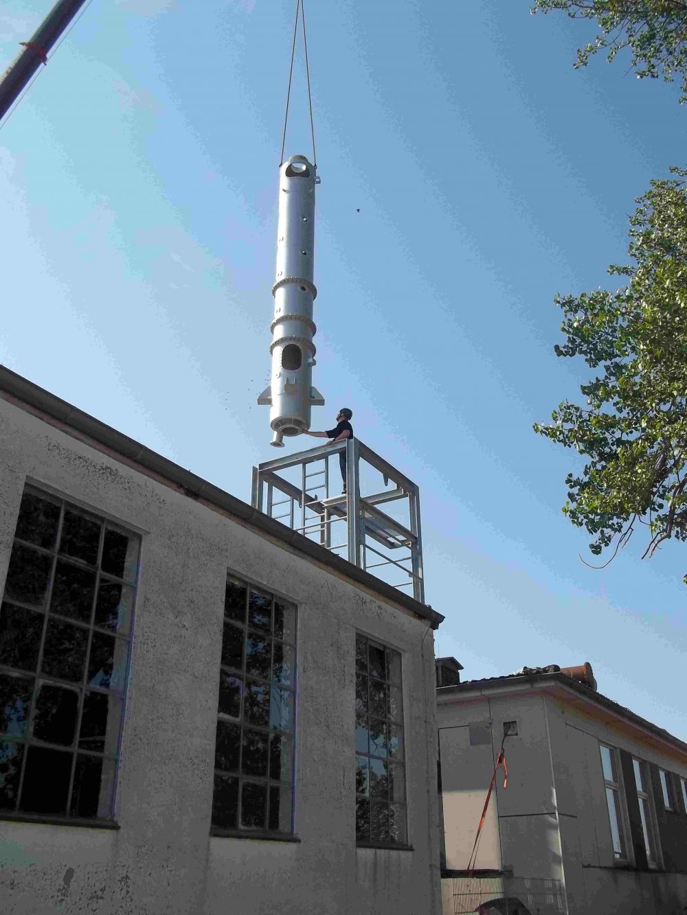 Auf einem der Zweckbauten aus den vorigen Bildern sitzt nun ein Metallgerüst. Darüber schwebt von einem Kran getragen ein großer, zylinderförmiger Turm. Der Himmel ist blau.