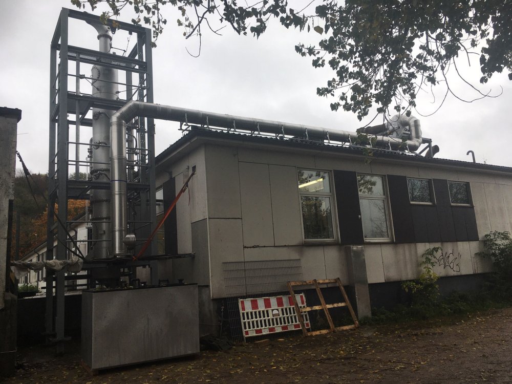 Ein schmuckloser, eingeschossiger Betonbau vor bewölktem Himmel. Neben dem Bau ist eine Turmkonstruktion zu sehen von der aus ein großes Rohr zum Teil über das Dach des Gebäudes verläuft und dann aus dem Sichtfeld verschwindet.