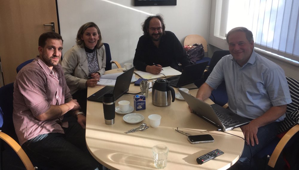 Vier Personen sitzen im Halbrund an einem Besprechungstisch, auf dem Tisch vor ihnen aufgeklappte Laptops, Notizbücher, Tassen, Kaffeekannen und ähnliche Besprechungsutensilien. Alle vier blicken in die Kamera und lächeln.