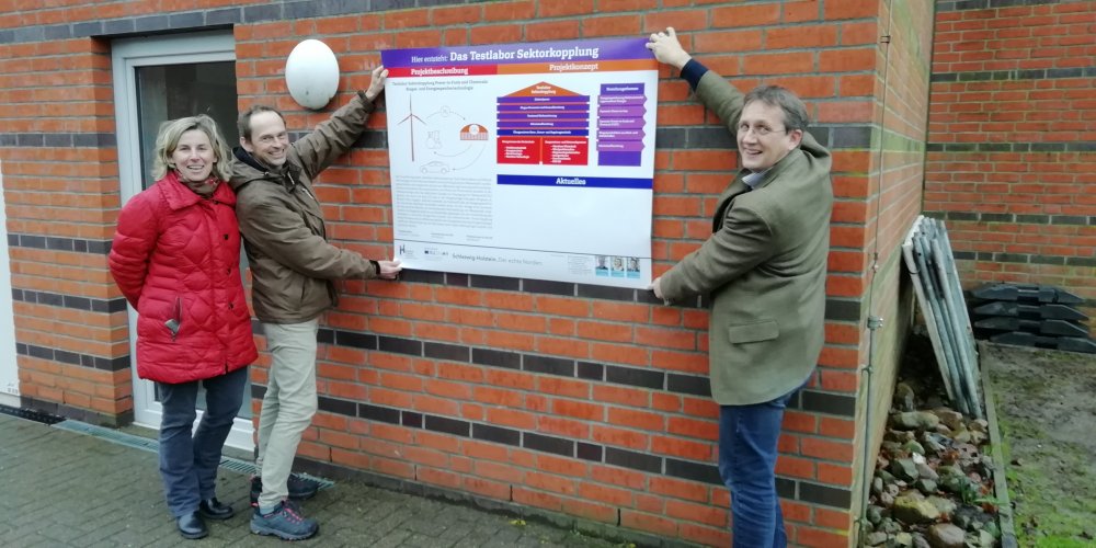 Zwei Männer halten ein Plakat auf dem die Farben weiß, lila und rot dominieren an eine Backsteinwand. Sie stehen links und rechts vom Plakat, noch weiter links steht eine Frau. Alle drei lachen.