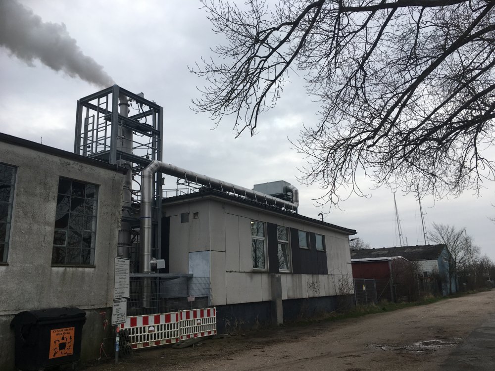 Zwei unverputzte einschossige Gebäude. An der Wand verläuft ein Metallroihr, zwischen den Gebäuden steht ein Metallrohr, das von einem Gerüst umgeben ist. Aus dem Rohr raucht oder qualmt es.