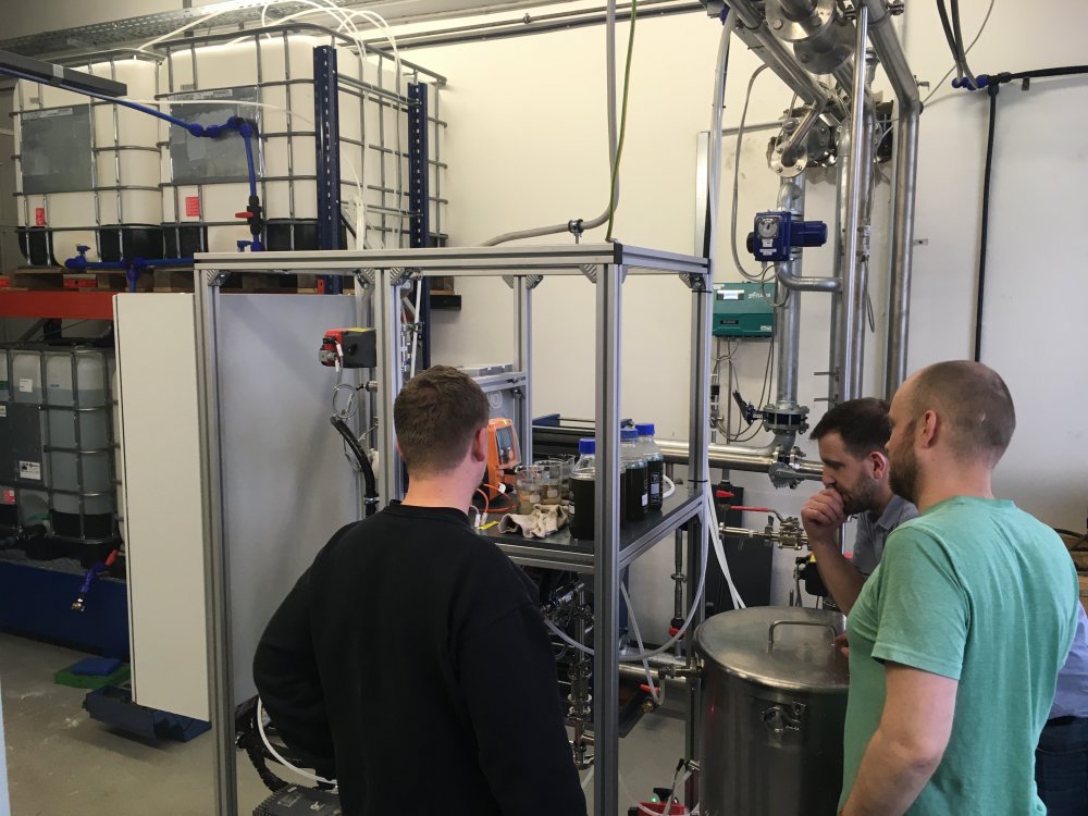 Drei Männer betrachten einen Aufbau. An und auf dem Aufbau befinden sich Messgeräte und Flaschen mit einer dunklen Flüssigkeit.