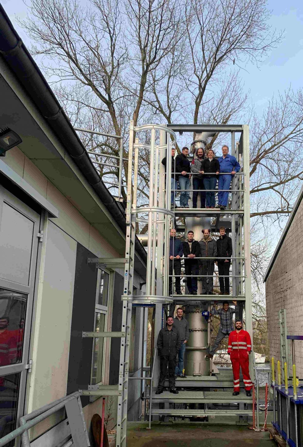 Metallrohr mit begehbarem Gerüst drumherum. Auf diesem Gerüst haben sich über drei Ebenen Menschen aufgestellt und lachen für die Kamera. Es ist ein sonniger Wintertag, der Himmel ist blau, die Bäume kahl.