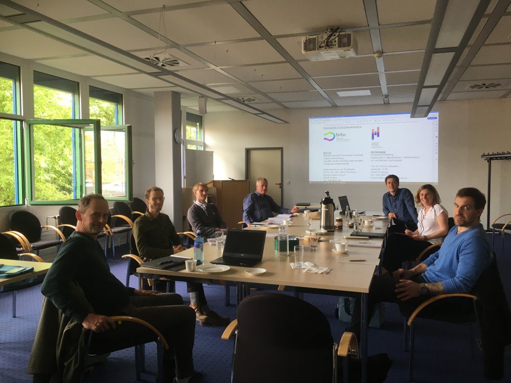 Eine Gruppe von Menschen sitzt um einen Besprechungstisch, hinter ihnen wird etwas an die Wand projiziert. Links im Bild sind durch ein Fenster sonnenbeschienene Bäume zu erkennen.