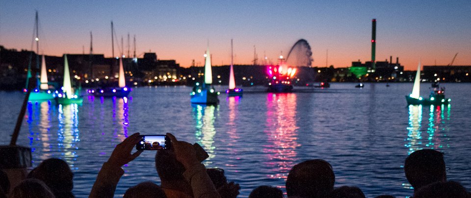 beleuchtete Boote auf dem Wasser, im Vordergrunf Publikum und die Szene in klein auf einem Smartphonedisplay
