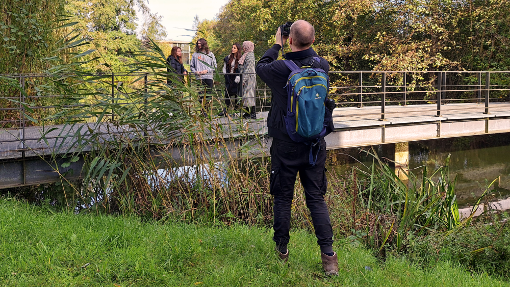 Ein junger Fotograf steht auf einer Wiese und fotografiert vier Studierende, die auf einer Brücke posieren.