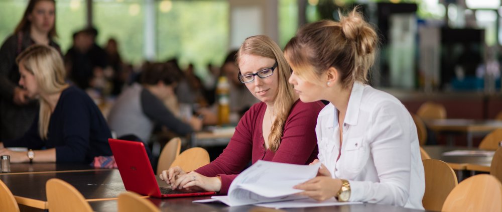 Zwei Studentinnen arbeiten gemeinsam an einem Laptop, eine blättert außerdem in Unterlagen.