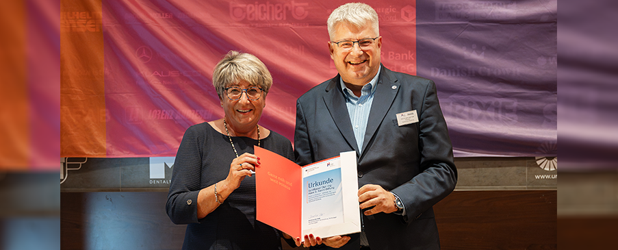 Eine Frau und ein Mann, beide fortgeschrittenen Alters und in Anzug bzw. Kostüm, stehen vor einer bunten Flagge und halten lächelnd eine Urkunde in die Kamera.