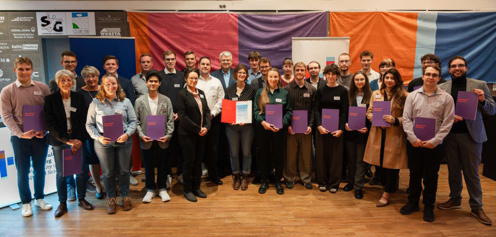 Eine Gruppe von jungen Menschen steht auf einer Bühne, ein älterer Mann überreicht ihnen nach und nach Urkunden. Hinter ihnen an der Wand stehen Aufsteller mit den Logos des Deutschlandstipendiums, der Hochschule Flensburg und der Europa-Universität Flensburg.