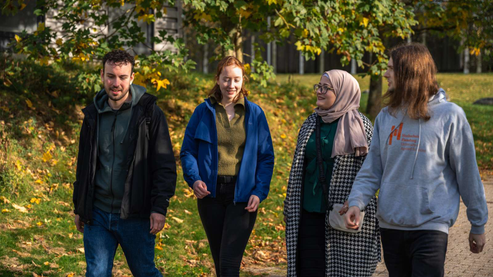 Vier junge Menschen laufen im Sonnenschein über einen Gehweg, im Hintergrund eine grüne Wiese und Bäume. Eine von ihnen trägt ein Kopftuch, ein anderer trägt einen grauen Kapuzenpulli mit dem Logo der Hochschule Flensburg.