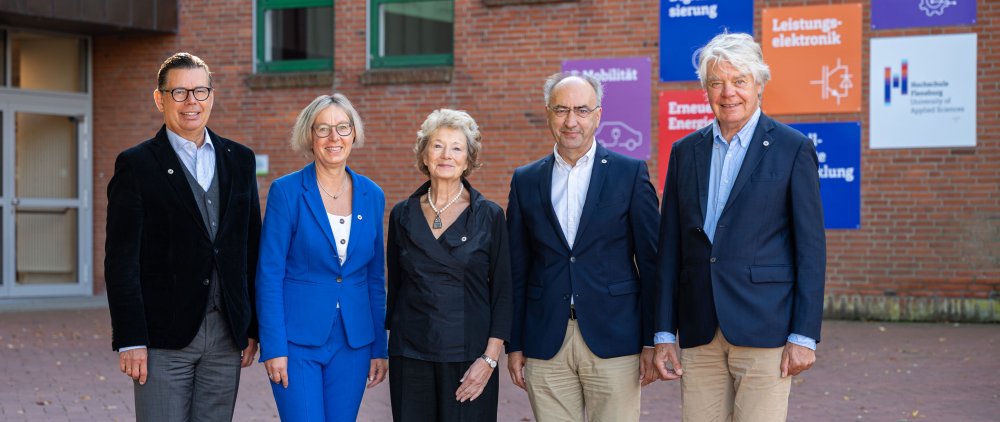 Drei Männer und zwei Frauen posieren vor einem Hochschulgebäude für ein Gruppenbild. Im Hintergrund u.a. Kacheln in Hochcshulfarben und das Hochschul-Logo.