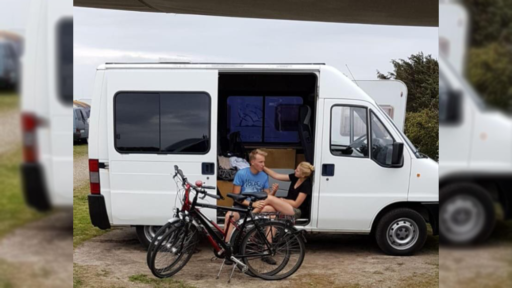 Ein junges Paar sitzt in der offenen Tür eines weißen Camper-Vans und unterhält sich miteinander, vor ihnen stehen zwei Fahrräder.