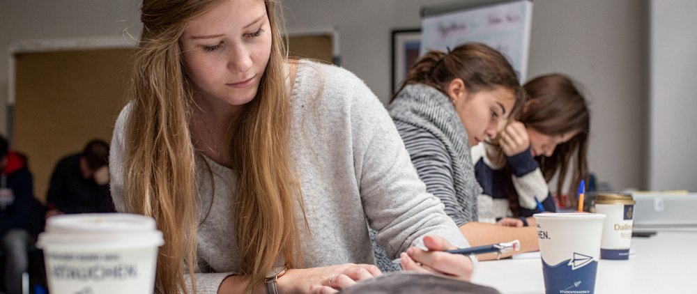 Studentin mit Stift in fder Hand an einem Tisch sitzend, auf dem Tisch Kaffeebecjer und im Hintergrund zwei weitere Studentinnen, die über Unterlagen gebeugt sind und Stifte in den Händen halten.