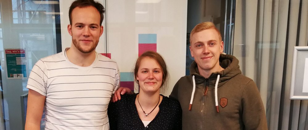 Mentorin und Mentoren des Sommersemester 2019: Eine Studentin und zwei Studenten posieren vor einer Glaswand, verdeckt ist auch das Hochschulloge zu erahnen.